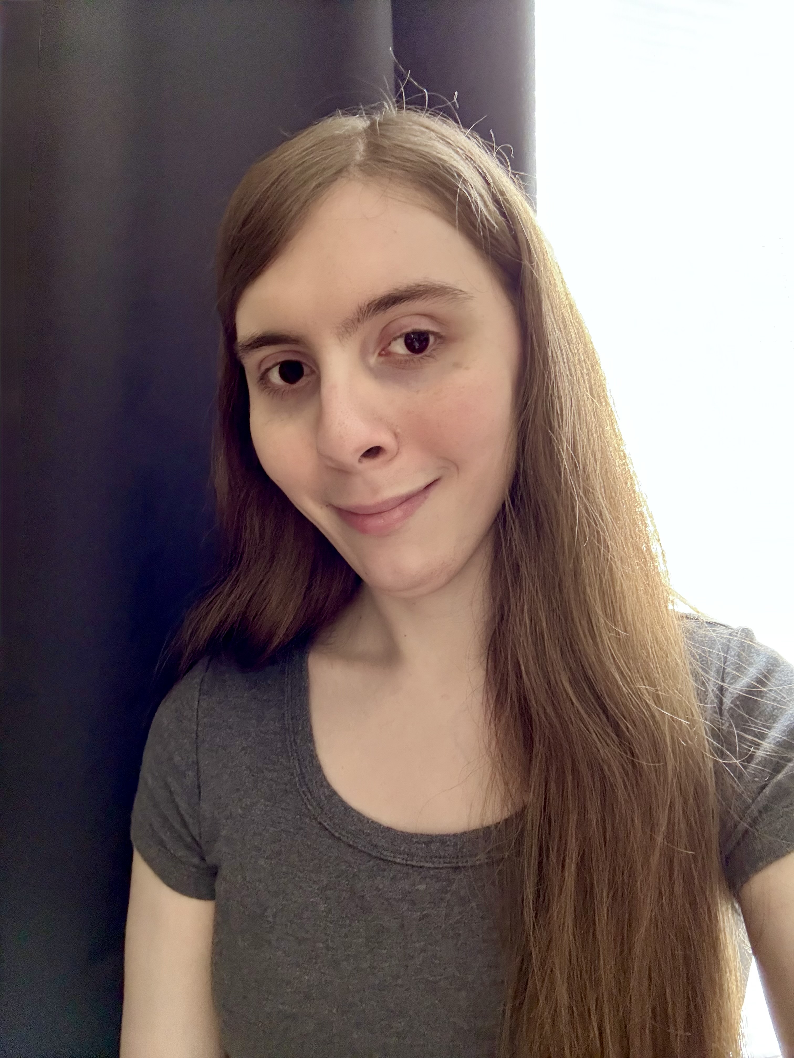 Photo of me! A selfie of a white trans woman with long dark blonde hair and brown eyes wearing a scoop-neck tee smiling and sitting in front of a window with dark curtains.